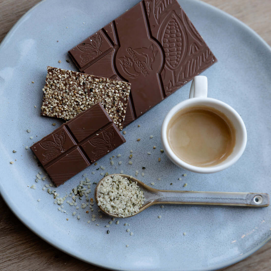 Flatlay mit Genussfux Bio-Hanf-Schokolade, bestreut mit gerösteten Hanfsamen, neben Espresso und Löffel voller geschälter Hanfsamen
