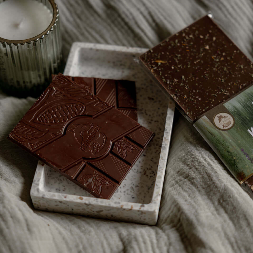 Handgegossene Bio-Zartbitterschokolade (54 % Kakao) mit getrockneten Wildgartenkräutern auf Terrazzo-Tablett im Flatlay mit Kerze und Leinenstoff