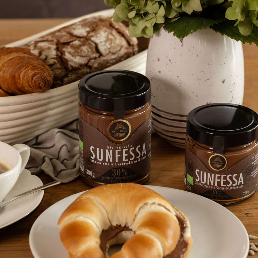 Brunch-Flatlay auf Holztisch mit zwei Gläsern Genussfux Sunfessa Schokocreme (30 % geröstete Bio-Sonnenblumenkerne), palmölfrei, neben Croissant und Kaffee