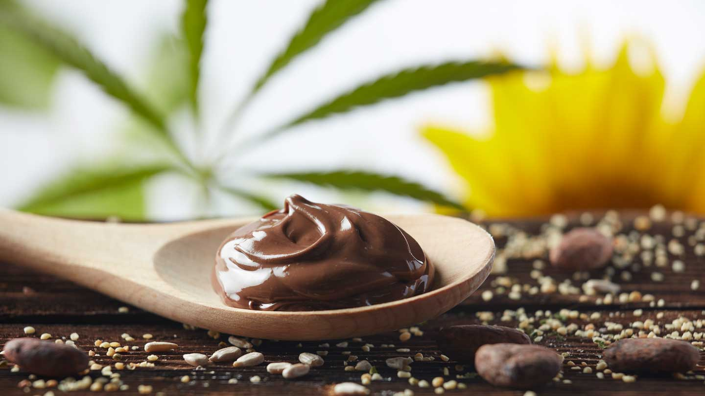 Nougatcreme liegt auf dem Holzlöffel und im Hintergrund sieht man ein Hanfblatt und eine Sonnenblume