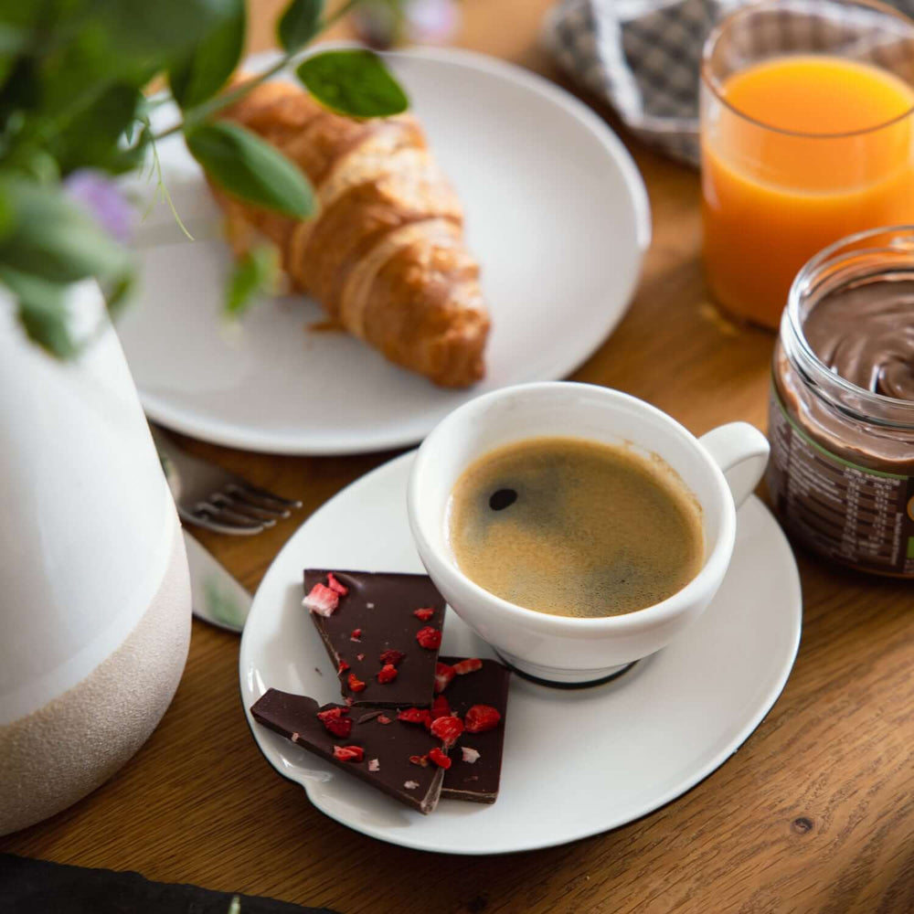 Frühstücks-Flatlay mit Tasse Kaffee, Croissant, Glas Orangensaft und Stücken Genussfux Bio-Erdbeer-Chili-Zartbitterschokolade