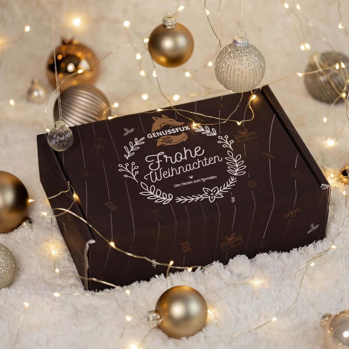 Genussfux Geschenkbox mit weißem “Frohe Weihnachten”-Kranzdesign auf Kunstfell, umgeben von Lichterkette und Weihnachtskugeln