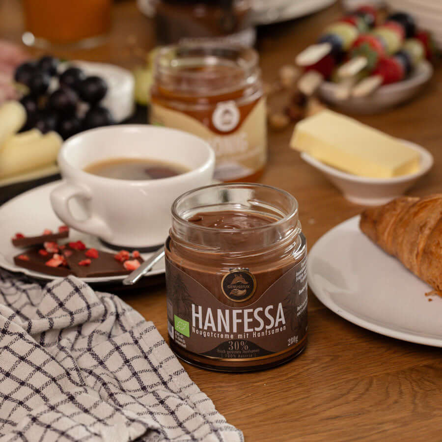 Brunch-Flatlay auf Holztisch mit Genussfux Hanfessa Nougatcreme 280 g (30 % geröstete Hanfsamen), palmölfrei, neben Kaffee, Croissant und Honig