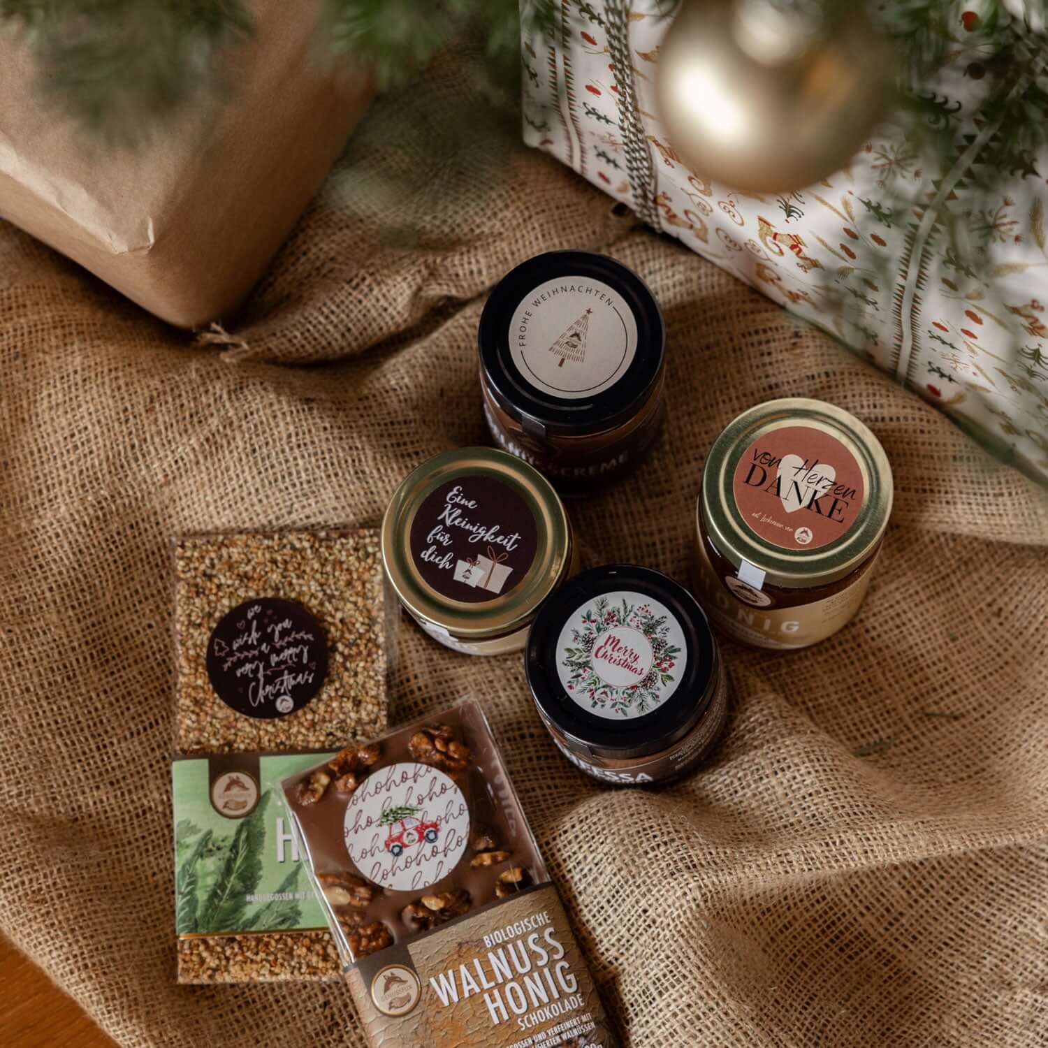 Genussfux Weihnachts-Geschenkset mit vier Nougat-Cremen, Bio-Walnuss-Honig-Schokolade und kostenlosen Weihnachts-Stickern auf Jute-Hintergrund
